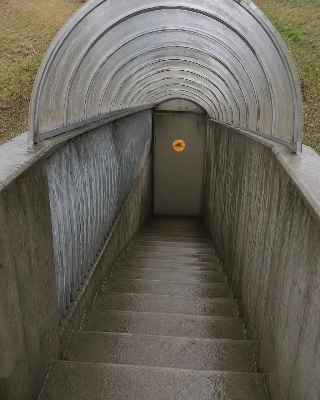 Looking down the covered concrete stair on a wet ordinary day: corrugated galvanized hood above, thirteen steps down to a smooth flush door carrying only a small lit porthole