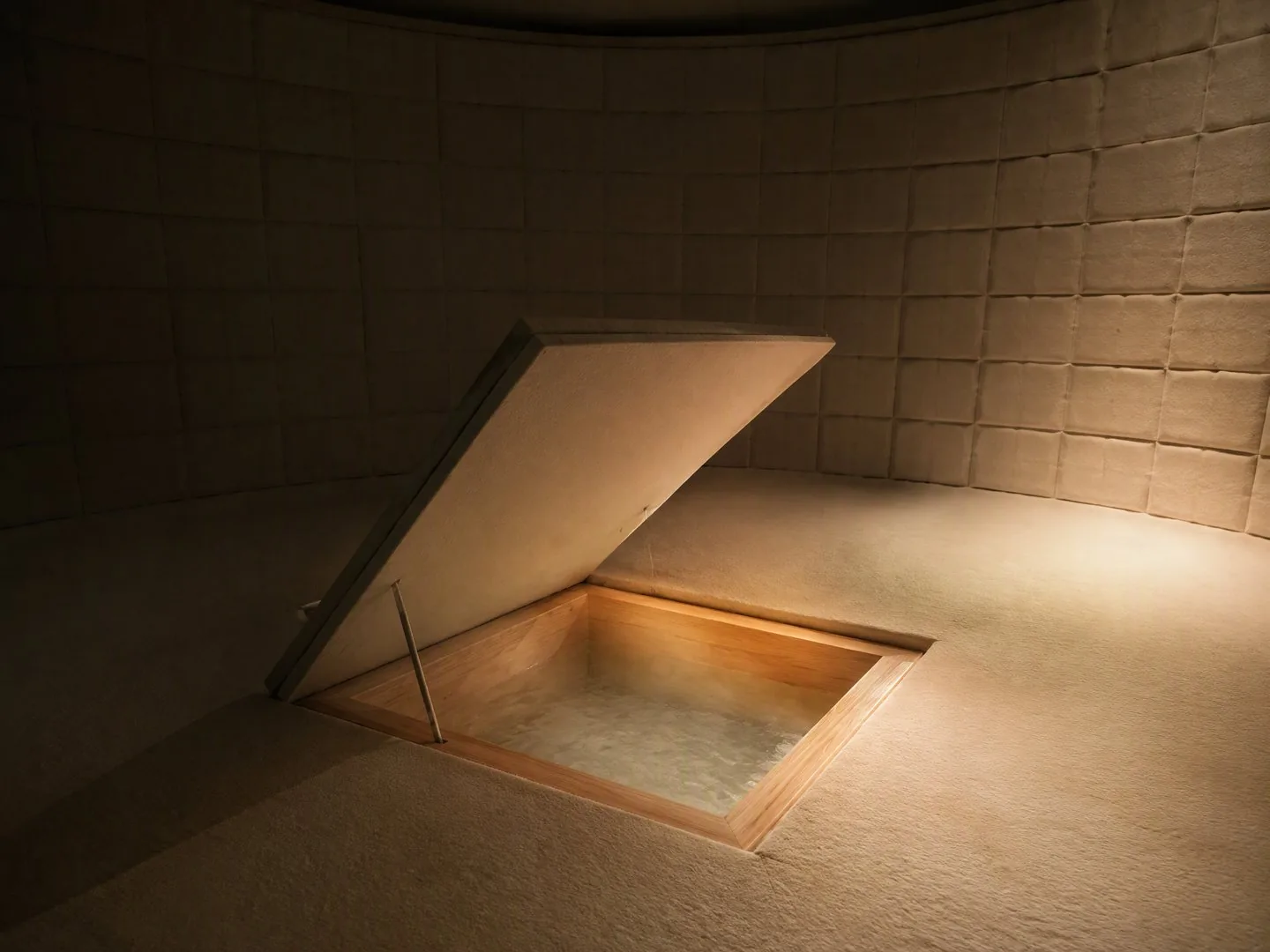 One flush floor panel propped open revealing the sunken hinoki bath, steam rising, warm single-source light, quilted walls behind